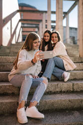 Smiling teenage girl taking selfie with friends while sitting on steps in city - GRCF00641
