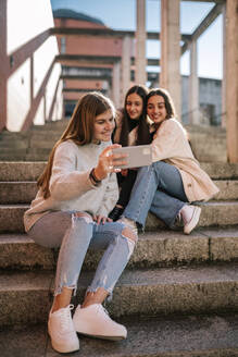 Lächelndes Teenager-Mädchen, das ein Selfie mit Freunden macht, während es auf einer Treppe in der Stadt sitzt - GRCF00641