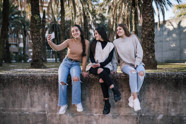 Lächelndes Teenager-Mädchen, das ein Selfie mit Freunden macht, während es auf einer Stützmauer im Park sitzt - GRCF00652