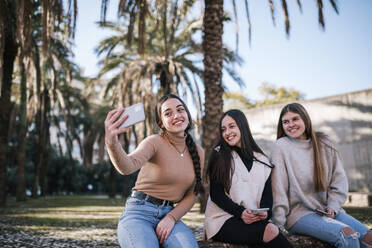 Lächelndes Teenager-Mädchen, das ein Selfie mit Freunden macht, während es gegen Bäume im Park sitzt - GRCF00653