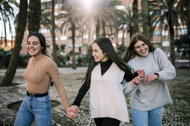 Cheerful female friends holding hands while walking on footpath in park - GRCF00658