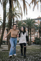 Fröhliche Freundinnen halten sich an den Händen, während sie auf einem Fußweg im Park laufen - GRCF00662