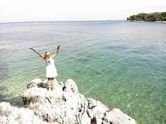 Frau mit erhobenen Armen und Blick auf das Meer, während sie an einem sonnigen Tag auf einem Felsen steht - PSIF00416