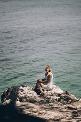 Ältere Frau sitzt auf einem Felsen am Strand vor einer Meereslandschaft an einem sonnigen Tag - PSIF00432