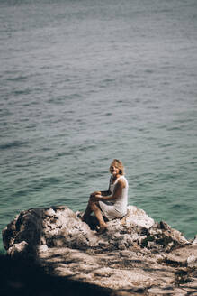 Ältere Frau sitzt auf einem Felsen am Strand vor einer Meereslandschaft an einem sonnigen Tag - PSIF00432