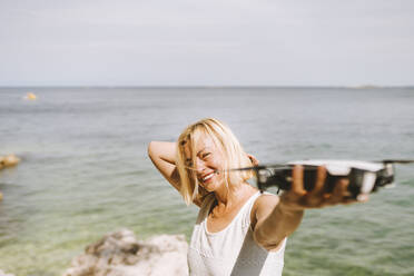 Smiling woman holding drone while standing against sea - PSIF00436