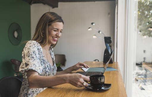 Gut gelaunte Unternehmerin mit Kaffeetasse auf dem Tisch und Laptop in einem Café - DSIF00282