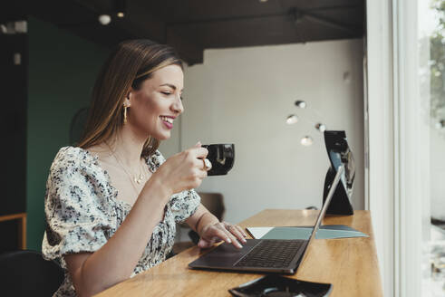 Geschäftsfrau, die eine Kaffeetasse in der Hand hält, während sie an einem Laptop auf einem Tisch im Restaurant arbeitet - DSIF00284