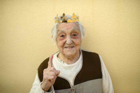 Close-up of smiling senior woman wearing paper crown gesturing against wall - RAEF02422