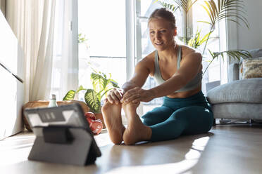 Woman with digital tablet stretching while exercising at home - JSRF01270