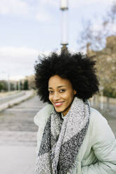 Smiling Afro woman with hands in pockets standing outdoors during winter - XLGF01030