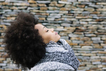 Young woman with eyes closed in warm clothing by stone wall - XLGF01037