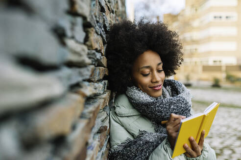 Smiling Afro woman writing in book while leaning on stone wall during winter - XLGF01038