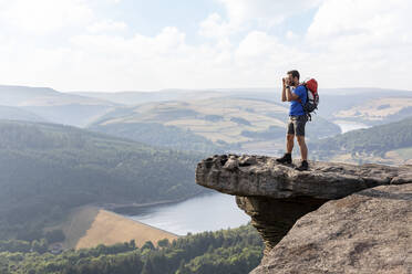 Young male hiker looking through binoculars while standing on cliff during vacations - WPEF03971