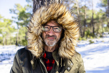 Lächelnder Mann mit Wintermantel, der im Winter im Wald steht und starrt - DLTSF01551