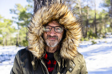 Smiling man wearing winter coat staring while standing in forest during winter - DLTSF01551