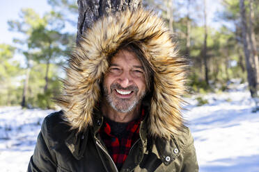 Mature man wearing fur hood smiling while standing in forest - DLTSF01552