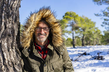 Mature man smiling while leaning on tree on forest - DLTSF01554