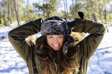 Smiling woman wearing fur cap standing with hands behind head at forest - DLTSF01556