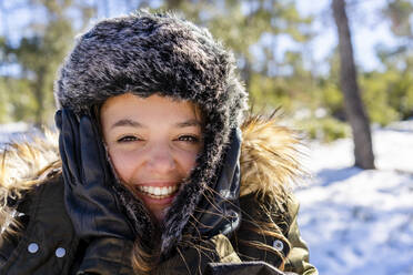 Cheerful woman wearing fur cap smiling while standing on forest during winter - DLTSF01557