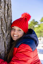 Junge Frau mit roter Strickmütze, die sich lächelnd an einen im Wald stehenden Baum schmiegt - DLTSF01562