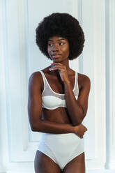 Afro young model with hand on chin in white lingerie looking away against wall - OIPF00195