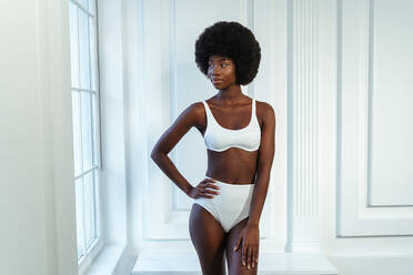 Afro young fashion model with hand on hip in white lingerie looking away against wall - OIPF00201