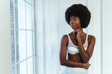 Afro weibliches Modell in Dessous mit Hand am Kinn schaut durch Fenster gegen weiße Wand - OIPF00204