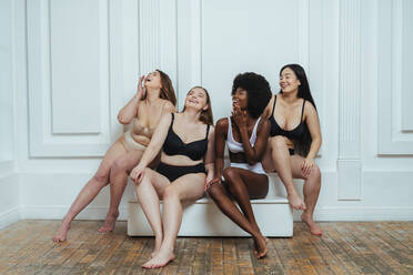 Multi-ethnische Gruppe von weiblichen Modellen in Dessous, die lachend an einer weißen Wand sitzen - OIPF00217
