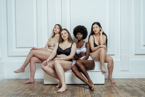 Confident multi-ethnic group of models in lingerie sitting against white wall - OIPF00219