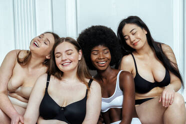 Glückliche multiethnische Gruppe von Modellen in Dessous mit geschlossenen Augen, die an einer weißen Wand sitzen - OIPF00221