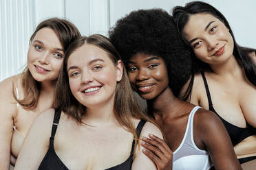 Lächelnde multiethnische Gruppe von Model-Freundinnen in Dessous, die zusammen vor einer weißen Wand sitzen - OIPF00222