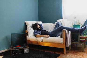 Bärtiger Mann hört Musik über einen Plattenspieler, während er zu Hause auf dem Sofa liegt - MRRF00842