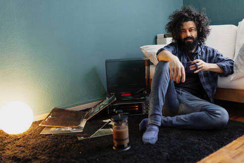 Bärtiger Mann mit wirrem Haar, der eine Kaffeetasse in der Hand hält, während er am Plattenspieler im Wohnzimmer sitzt - MRRF00846