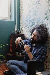 Hipster man holding camera while sitting cross legged against wall at home - MRRF00847