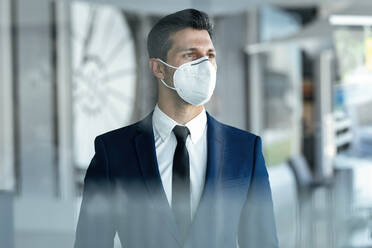 Businessman wearing protective face mask looking away while standing in office - JSRF01284