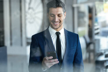 Smiling entrepreneur wearing suit using mobile phone while standing in office - JSRF01285