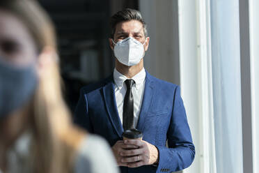Businessman wearing protective face mask holding coffee while standing in office - JSRF01295
