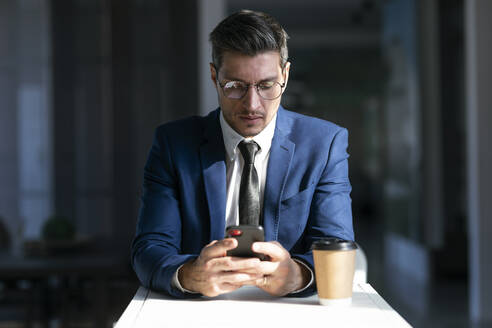 Unternehmer mit Brille, der ein Mobiltelefon benutzt, während er in der Cafeteria im Büro sitzt - JSRF01297