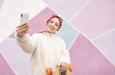 Junge Frau mit Skateboard, die ein Selfie macht, während sie vor einer mehrfarbigen Wand steht - JCCMF00927