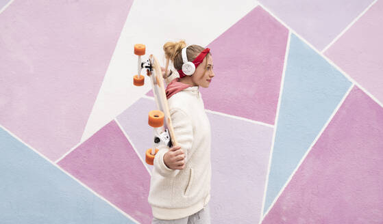 Junge Frau mit Kopfhörern, die ein Skateboard auf den Schultern trägt, während sie gegen eine bunte Wand läuft - JCCMF00930