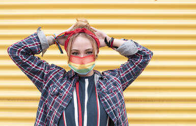 Young woman wearing colorful protective face mask standing with hands behind head against yellow wall - JCCMF00948