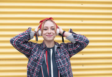 Woman wearing headphones smiling while standing against yellow wall - JCCMF00949