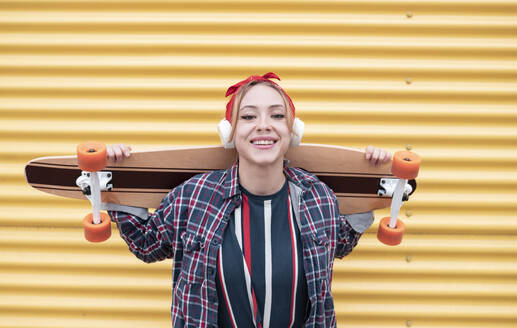 Lächelnde Frau mit Kopfhörern, die ein Skateboard auf den Schultern trägt und an einer gelben Wand steht - JCCMF00950