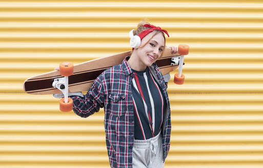 Junge Frau lächelt, während sie ein Skateboard auf der Schulter gegen eine gelbe Wand trägt - JCCMF00951