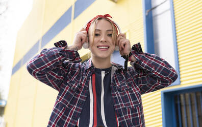 Young woman smiling while adjusting headphones standing outdoors - JCCMF00952