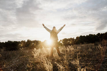 Senior male hiker with arms raised standing in filed during sunset - AFVF08112