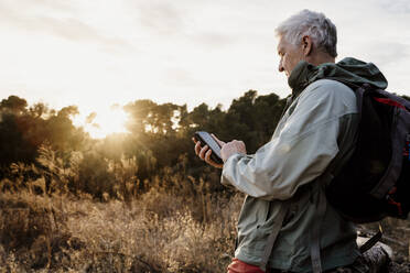 Älterer männlicher Wanderer, der während des Sonnenuntergangs auf einem landwirtschaftlichen Feld sein Smartphone benutzt - AFVF08118