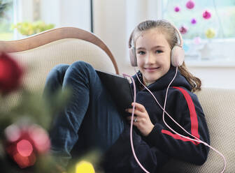 Lächelndes Mädchen hört Musik auf einem digitalen Tablet, während sie zu Hause auf dem Sofa sitzt, während Weihnachten - DIKF00545