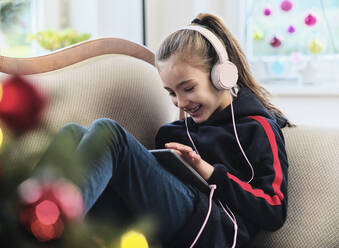 Cheerful girl listening radio on digital tablet while sitting on sofa in living room during holiday - DIKF00546